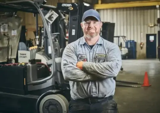 A smiling forklift mechanic with safety glasses and a cap poses with his arms crossed in a forklift repair shop in Thompson Lift Truck Alabama