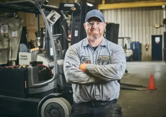 A professional forklift mechanic in Georgia stands with his arms crossed in front of a Crown forklift inside a repair and service bay